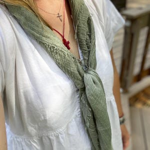 Op de afbeelding: Een lichtgroene sjaal met een gerafelde rand is over een witte jurk gedrapeerd. Een zilveren ketting met een kruisvormige hanger en een rode kralen kruisketting zijn zichtbaar.