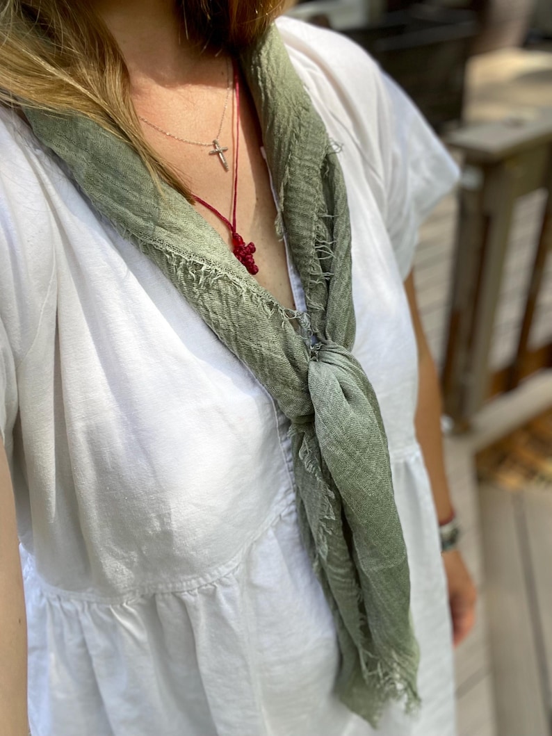 Op de afbeelding: Een lichtgroene sjaal met een gerafelde rand is over een witte jurk gedrapeerd. Een zilveren ketting met een kruisvormige hanger en een rode kralen kruisketting zijn zichtbaar.