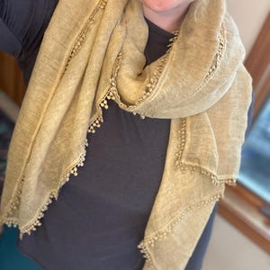 May include: A light tan scarf with a textured, semi-sheer fabric and a delicate, small pom-pom trim along the edges. The scarf is draped around the neck, showcasing the woven material and the consistent decorative fringe detail throughout.