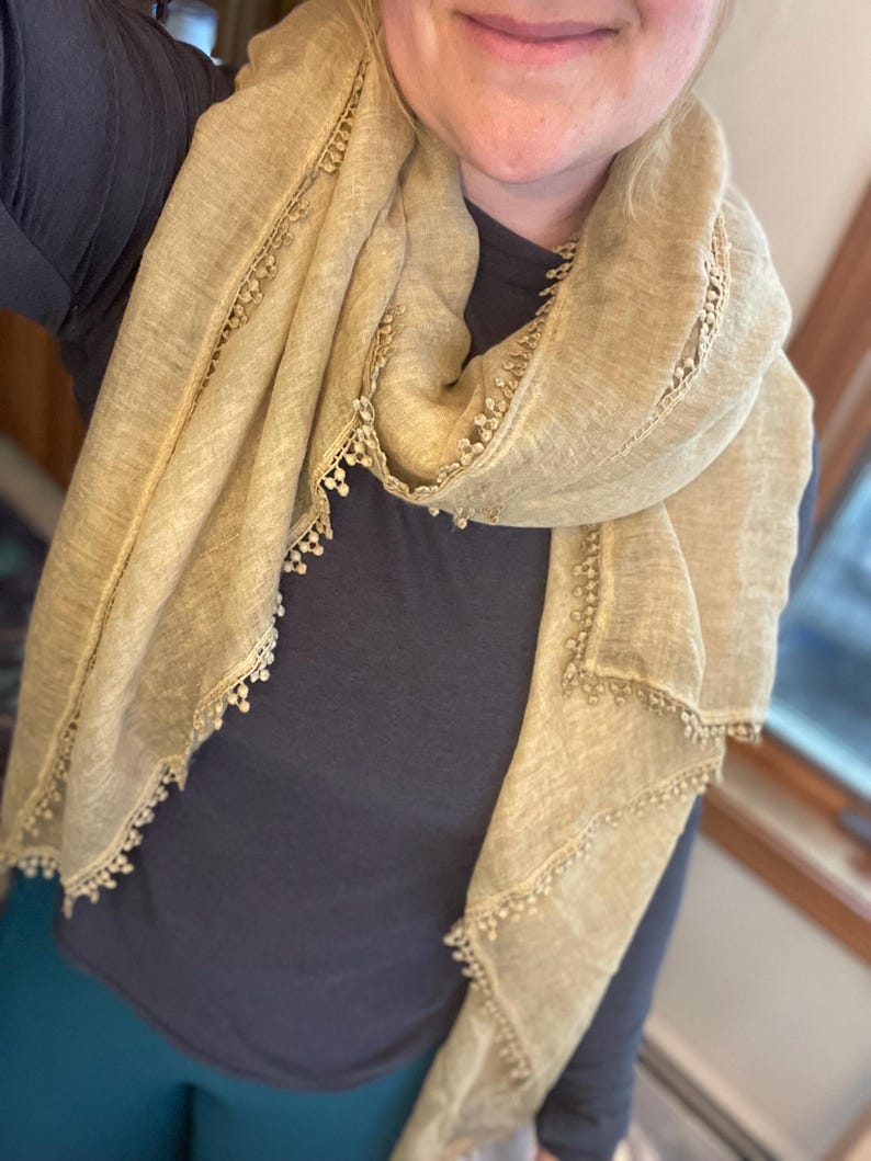 May include: A light tan scarf with a textured, semi-sheer fabric and a delicate, small pom-pom trim along the edges. The scarf is draped around the neck, showcasing the woven material and the consistent decorative fringe detail throughout.