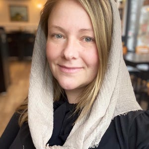 Op de afbeelding: Een vrouw met blond haar draagt een lichtbeige sjaal over haar hoofd en om haar nek geknoopt. De sjaal heeft een textuur. Ze draagt een zwarte top. De achtergrond is wazig, wat wijst op een binnenomgeving.