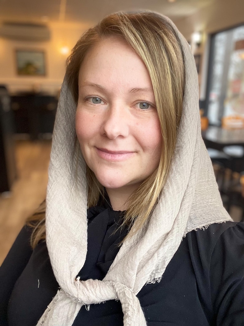 Op de afbeelding: Een vrouw met blond haar draagt een lichtbeige sjaal over haar hoofd en om haar nek geknoopt. De sjaal heeft een textuur. Ze draagt een zwarte top. De achtergrond is wazig, wat wijst op een binnenomgeving.