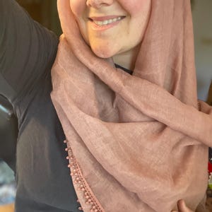 May include: A dusty rose-colored headscarf featuring a lightweight, textured fabric and a delicate trim along the edge adorned with small, matching spherical beads. The scarf is draped loosely around the head and shoulders, showcasing the soft pink hue.