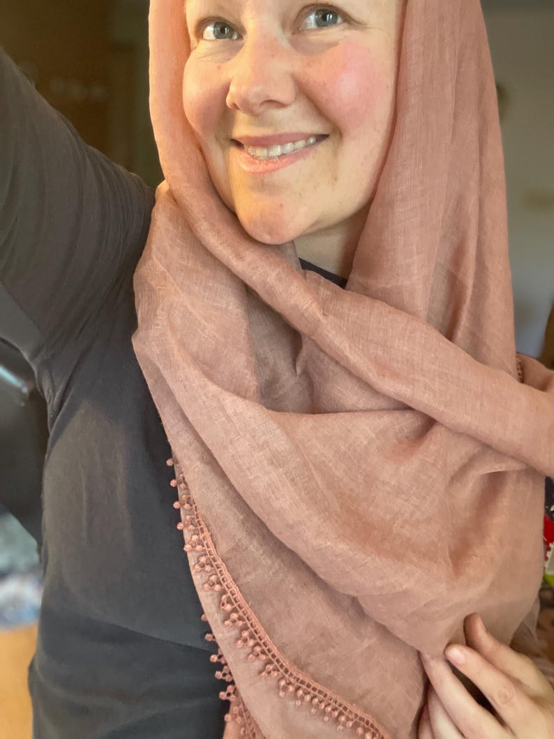 May include: A dusty rose-colored headscarf featuring a lightweight, textured fabric and a delicate trim along the edge adorned with small, matching spherical beads. The scarf is draped loosely around the head and shoulders, showcasing the soft pink hue.