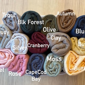 Op de afbeelding: Een verzameling opgerolde stoffen in verschillende kleuren, waaronder grijs, groen, bruin en blauw. De stoffen zijn gelabeld met kleurnamen zoals "Trout", "Blk Forest", "Autumn" en "Blues". De opstelling bevindt zich in een witte container.