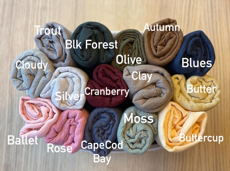 Op de afbeelding: Een verzameling opgerolde stoffen in verschillende kleuren, waaronder grijs, groen, bruin en blauw. De stoffen zijn gelabeld met kleurnamen zoals "Trout", "Blk Forest", "Autumn" en "Blues". De opstelling bevindt zich in een witte container.