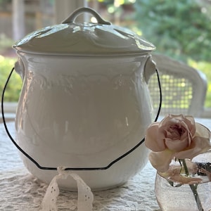 May include: A white ceramic lidded pot with a black wire handle and decorative floral detailing. A small glass with a pink rose is next to the pot. A white lace ribbon is in front of the pot.