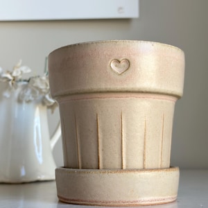 May include: A pale pink and beige ceramic planter with a heart design. The planter has a ribbed texture and a matching saucer.