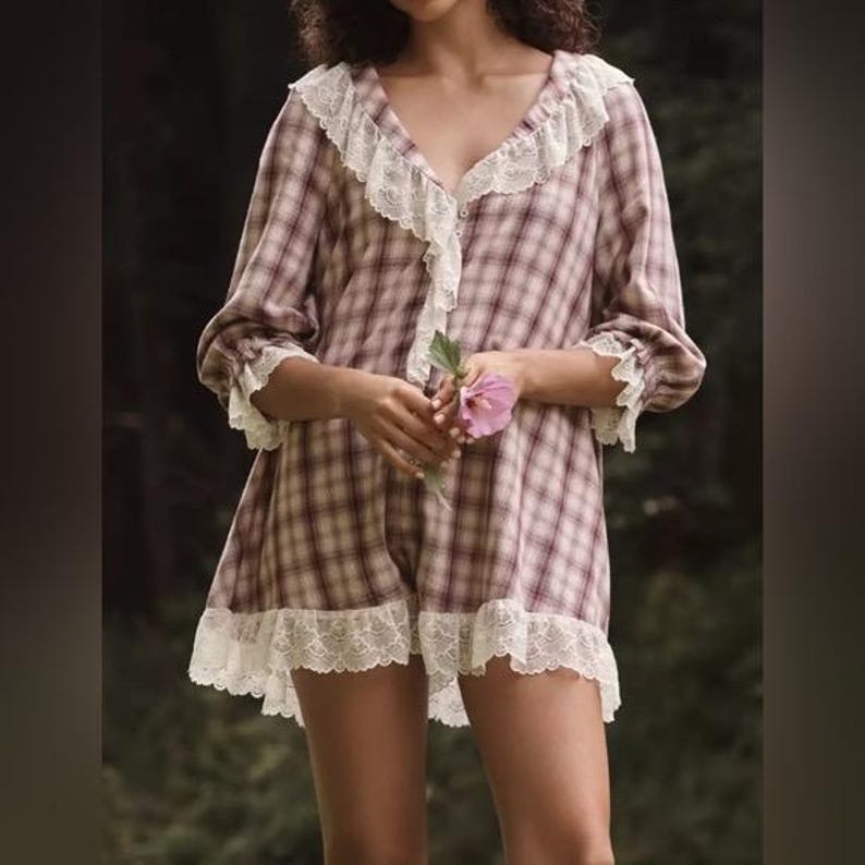 Op de afbeelding: Een geruite romper in tinten bordeauxrood en cr&egrave;me, afgewerkt met witte kant. De romper heeft een V-hals en korte mouwen, met kanten details op de kraag, manchetten en zoom. Een roze bloem wordt in het midden vastgehouden.