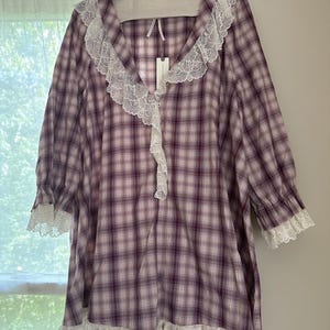 Op de afbeelding: Een paars en wit geruite nachtjapon met witte kanten afwerking. De nachtjapon heeft een V-hals met kanten afwerking en lange mouwen met kanten manchetten. De zoom heeft ook een witte kanten afwerking. Het kledingstuk hangt aan een grijze hanger.