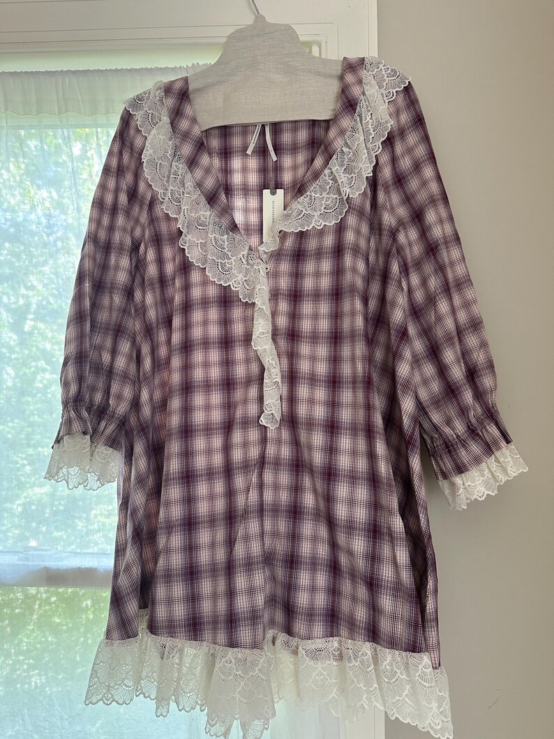 Op de afbeelding: Een paars en wit geruite nachtjapon met witte kanten afwerking. De nachtjapon heeft een V-hals met kanten afwerking en lange mouwen met kanten manchetten. De zoom heeft ook een witte kanten afwerking. Het kledingstuk hangt aan een grijze hanger.