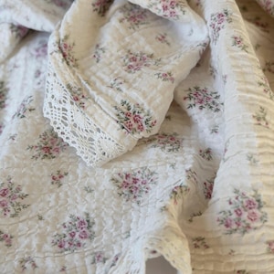 May include: Cream-colored fabric with a floral pattern of small pink and green flowers. The fabric has a textured, quilted appearance and is trimmed with delicate white lace. The overall aesthetic is soft and romantic.