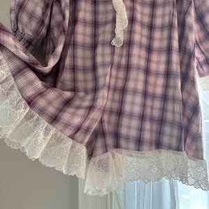 Op de afbeelding: Een vintage-stijl kledingstuk met lange mouwen in een paars en wit ruitpatroon. Het kledingstuk heeft witte kanten afwerking langs de zoom en halslijn. De mouwen zijn bij de manchetten gerimpeld.