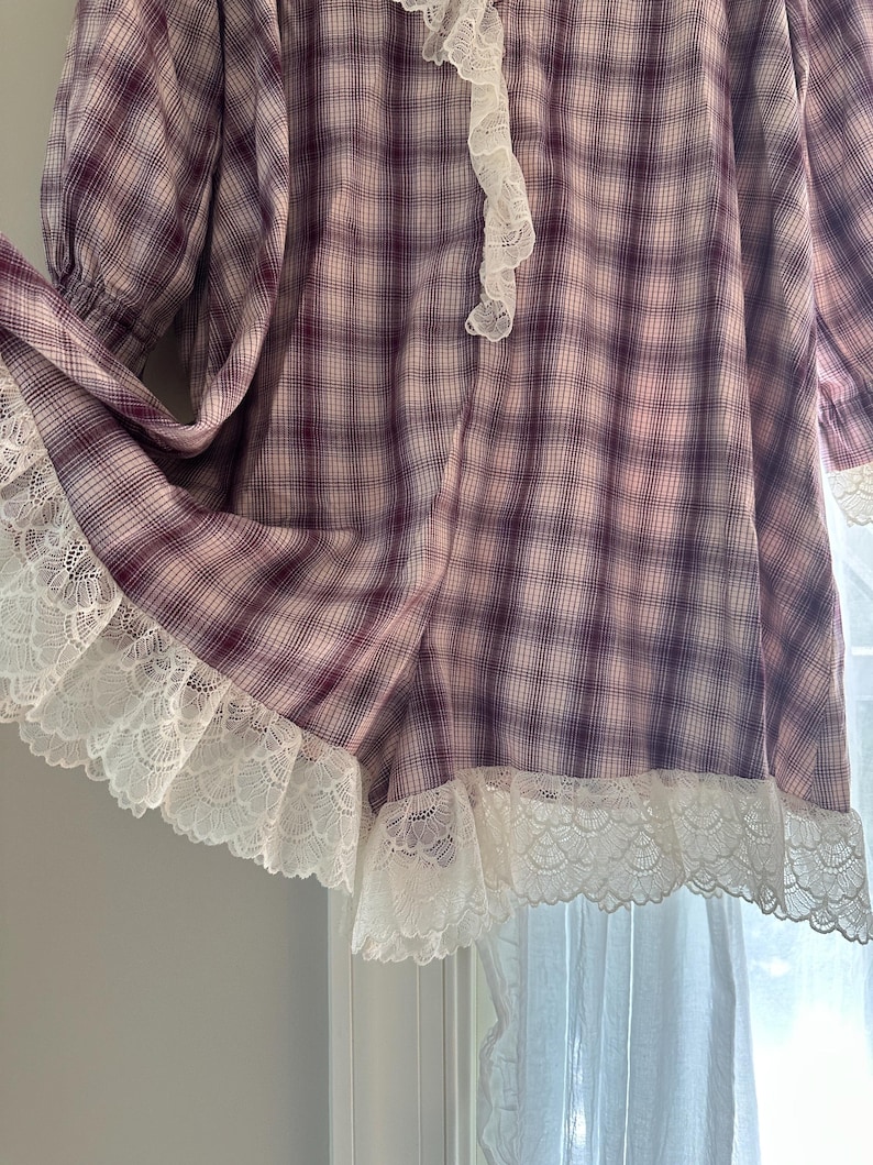 Op de afbeelding: Een vintage-stijl kledingstuk met lange mouwen in een paars en wit ruitpatroon. Het kledingstuk heeft witte kanten afwerking langs de zoom en halslijn. De mouwen zijn bij de manchetten gerimpeld.