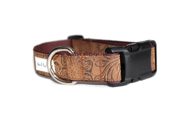 May include: A brown dog collar with a floral embossed pattern and a black plastic buckle. The collar has a silver D-ring.