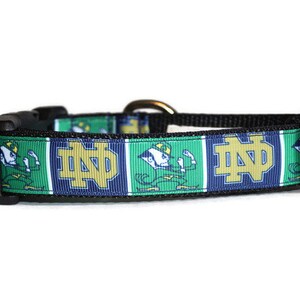 May include: A black dog collar with a green, blue, and gold Notre Dame Fighting Irish logo and mascot. The collar has a black buckle and a gold metal D-ring.