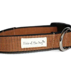 May include: A brown and black dog collar with a black buckle and a silver D-ring. The collar has a white label that says "Hair of the Dog" with paw prints.