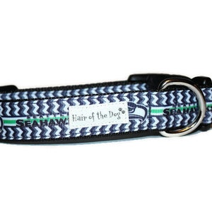 May include: A blue and white dog collar with a chevron pattern and the Seattle Seahawks logo. The collar has a silver buckle and the text "Hair of the Dog" is printed on the collar.
