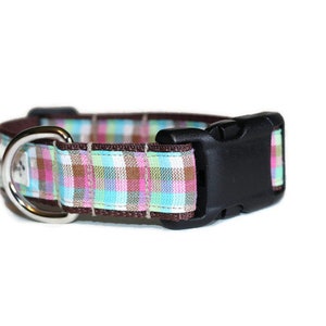May include: A brown dog collar with a plaid pattern in pink, blue, and green. The collar has a silver D-ring and a black plastic buckle.