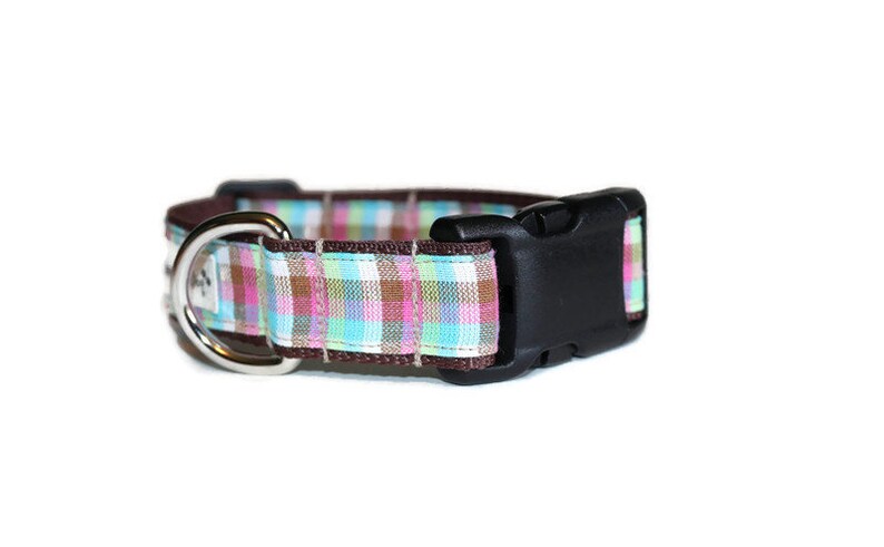 May include: A brown dog collar with a plaid pattern in pink, blue, and green. The collar has a silver D-ring and a black plastic buckle.