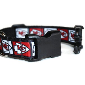 May include: A black dog collar with a red, white, and black patterned strap featuring the Kansas City Chiefs logo. The collar has a black plastic buckle and a silver D-ring.