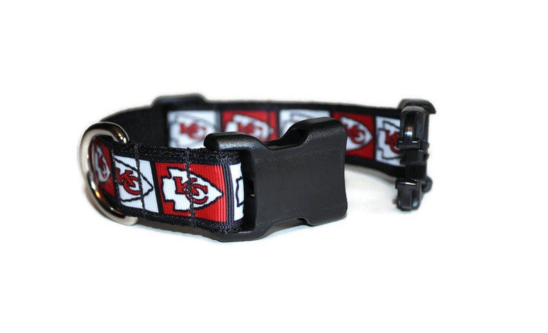 May include: A black dog collar with a red, white, and black patterned strap featuring the Kansas City Chiefs logo. The collar has a black plastic buckle and a silver D-ring.