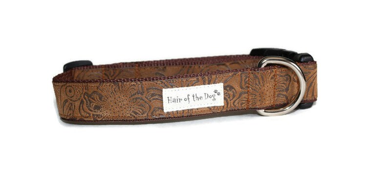 May include: A brown dog collar with a floral embossed design and a silver metal D-ring. The collar has a white label with the text "Hair of the Dog" printed on it.