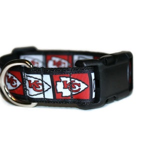 May include: A black dog collar with a red, white, and black patterned ribbon featuring the Kansas City Chiefs logo.