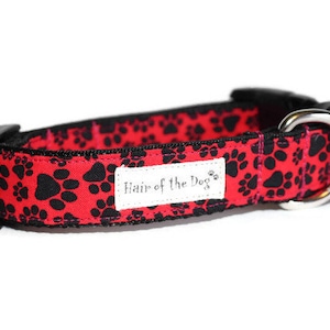 May include: Red dog collar with black paw print pattern and a silver metal D-ring. The collar has a black buckle and a white label that reads "Hair of the Dog."