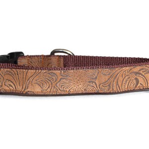 May include: A brown leather dog collar with a floral embossed pattern. The collar has a black buckle and a brown nylon strap.