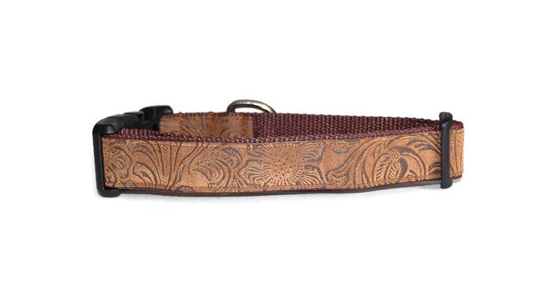 May include: A brown leather dog collar with a floral embossed pattern. The collar has a black buckle and a brown nylon strap.
