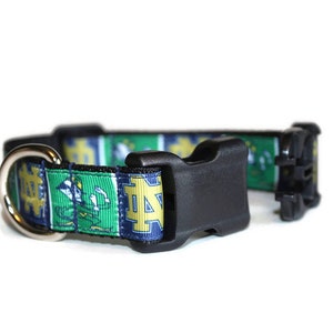 May include: A green, blue, and gold dog collar with a black buckle and a silver D-ring. The collar features a repeated pattern of the Notre Dame Fighting Irish logo.