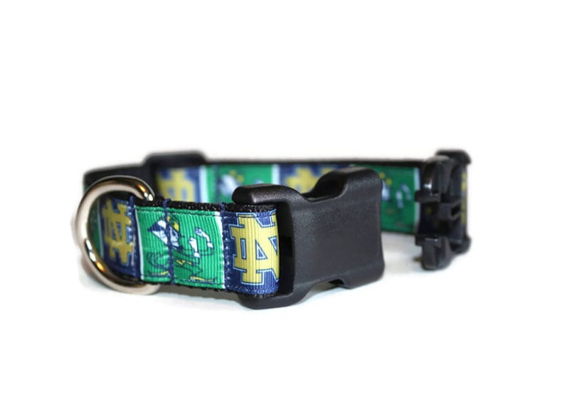 May include: A green, blue, and gold dog collar with a black buckle and a silver D-ring. The collar features a repeated pattern of the Notre Dame Fighting Irish logo.