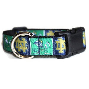 May include: A black and green dog collar with a silver D-ring and a black plastic buckle. The collar features a repeated pattern of the Notre Dame Fighting Irish logo and mascot, the leprechaun.