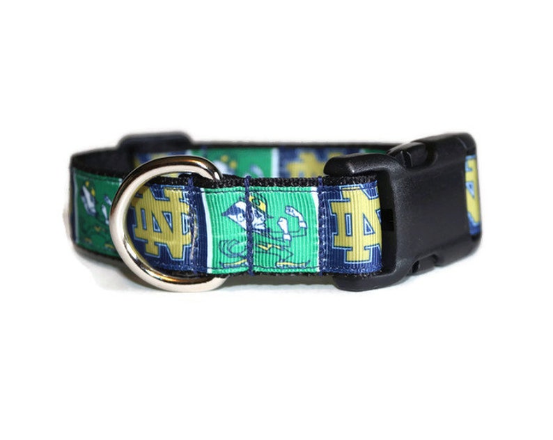 May include: A black and green dog collar with a silver D-ring and a black plastic buckle. The collar features a repeated pattern of the Notre Dame Fighting Irish logo and mascot, the leprechaun.
