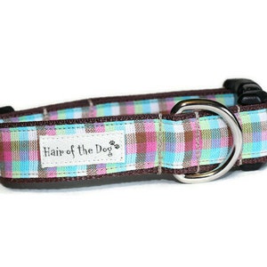 May include: A brown dog collar with a plaid pattern in pink, blue, and white. The collar has a silver D-ring and a white label that says "Hair of the Dog".