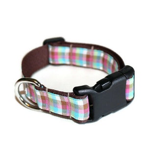 May include: A brown dog collar with a plaid pattern in pink, blue, and white. The collar has a black plastic buckle and a silver metal D-ring.