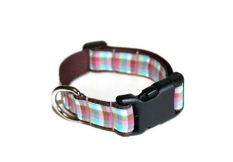 May include: A brown dog collar with a plaid pattern in pink, blue, and white. The collar has a black plastic buckle and a silver metal D-ring.