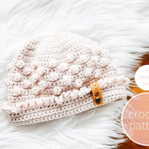 以下が含まれることがあります： 薄いピンク色のボブルステッチ模様のクロシェ編みのベビーハット。ハットはフリル付きの縁と、