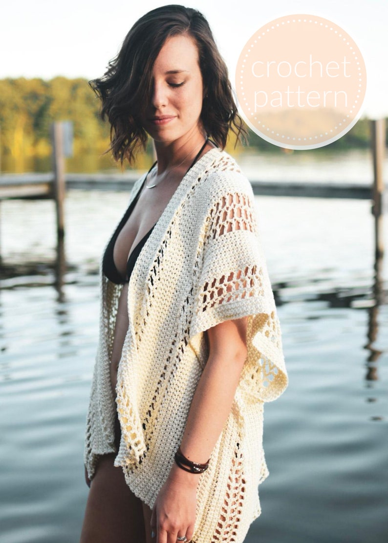May include: A white crochet cover-up with a lacy design. The cover-up has a V-neck and short sleeves. The model is wearing the cover-up over a black swimsuit.