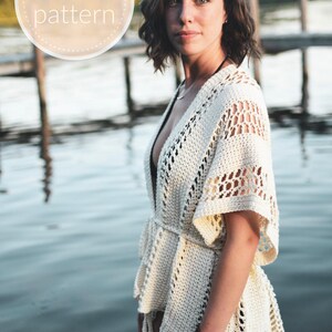 May include: A white crochet cover-up with a tie belt and a delicate openwork pattern. The cover-up is worn by a person standing near a body of water.