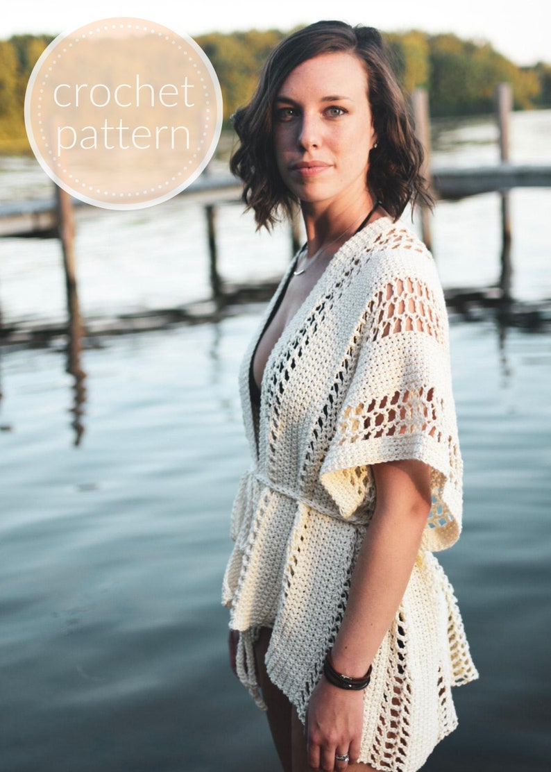 May include: A white crochet cover-up with a tie belt and a delicate openwork pattern. The cover-up is worn by a person standing near a body of water.