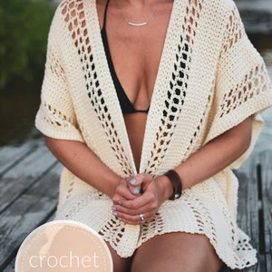May include: A white crochet cover-up with a delicate openwork pattern. The cover-up is worn over a black bikini top. The model is sitting on a wooden dock.
