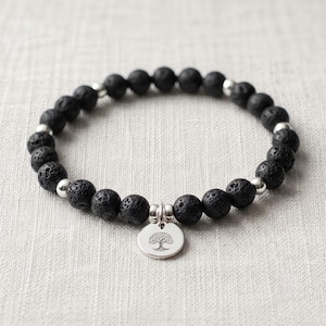 May include: A beaded bracelet featuring round, porous black lava stone beads interspersed with small, smooth silver-toned metal beads. A circular silver-toned metal charm hangs from the centre, engraved with a detailed tree of life design.