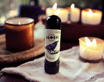 May include: A black glass bottle with a white label featuring a black raven and the text "Little Witches". The bottle is sitting on a white cloth with several lit candles in the background.