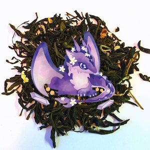 May include: A pile of loose leaf tea with a purple dragon sticker featuring white flowers and two crystal accents. The dragon has large wings and is surrounded by the dark green tea leaves.