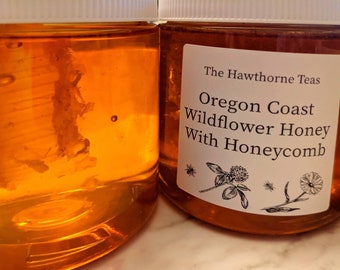May include: Two glass jars of honey with honeycomb. The jar on the right has a white label that reads "The Hawthorne Teas Oregon Coast Wildflower Honey With Honeycomb".