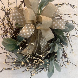 May include: A decorative wreath featuring a large burlap bow with white polka dots. The wreath includes green leaves, small white berries, and dark brown twigs, creating a natural and rustic aesthetic. The wreath is designed for home decor.
