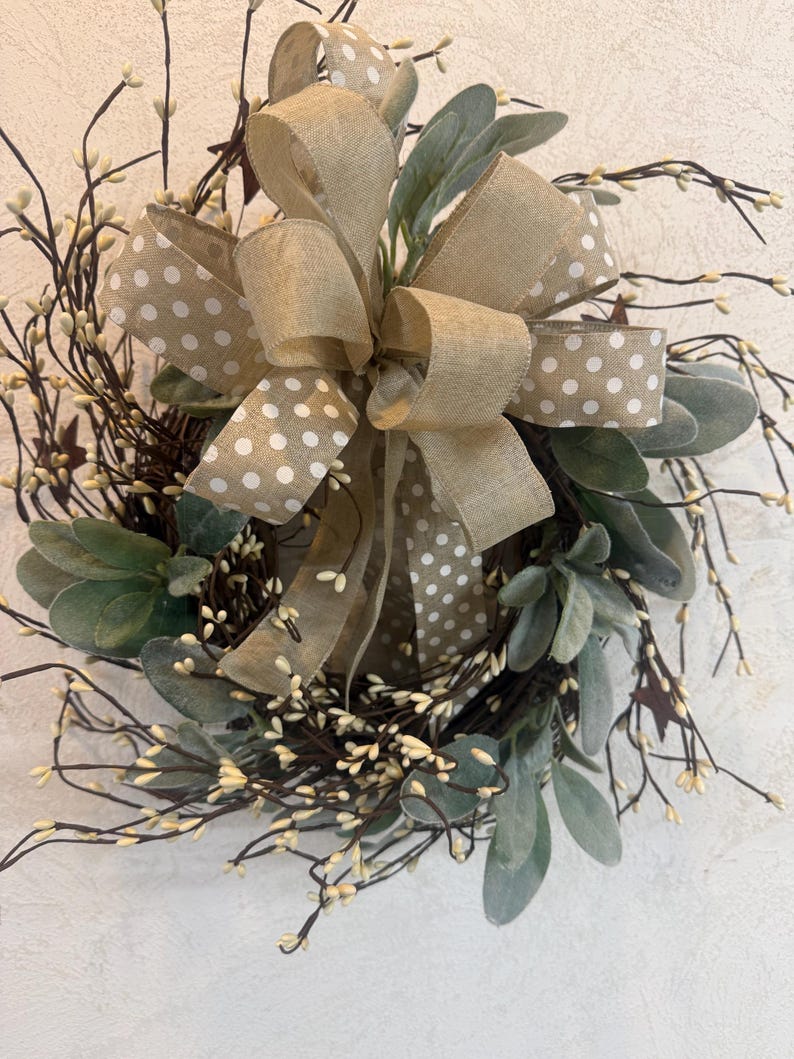 May include: A decorative wreath featuring a large burlap bow with white polka dots. The wreath includes green leaves, small white berries, and dark brown twigs, creating a natural and rustic aesthetic. The wreath is designed for home decor.