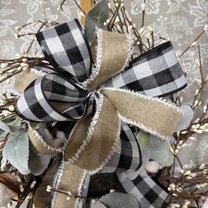 May include: A close-up of a decorative wreath featuring a large bow with black and white checkered ribbon and natural burlap ribbon. The wreath includes sprigs of white berries, green leaves, and cotton bolls, creating a rustic, farmhouse aesthetic.